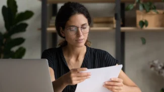 A woman is looking at a piece of paper in front of a laptop.