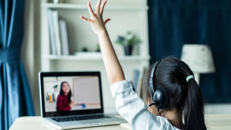 A girl is raising her hand while watching a video on her laptop.
