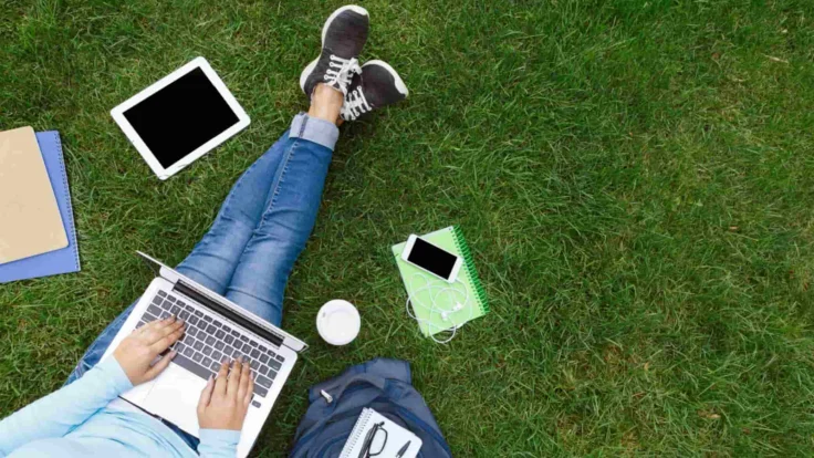 A woman is sitting on the grass with a laptop and other items.