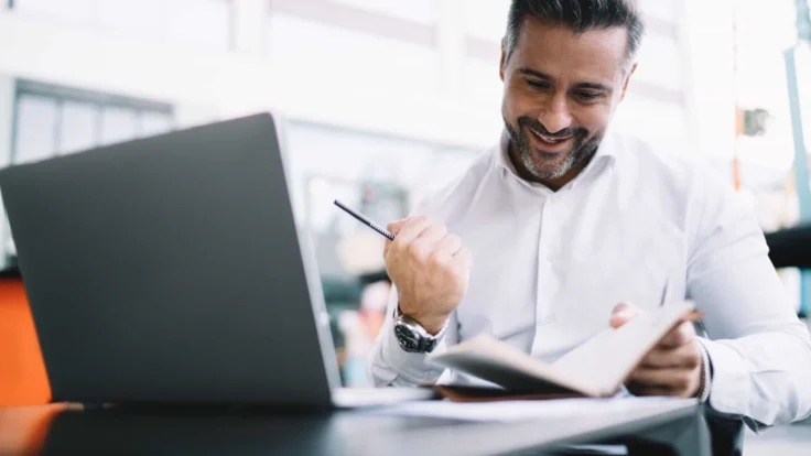 A man is sitting in front of a laptop and smiling.