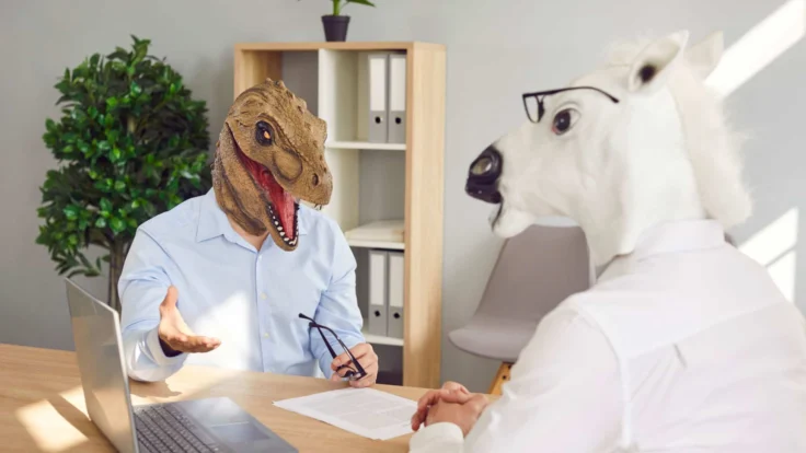 Two people sitting at a desk with a t - rex mask on their heads.