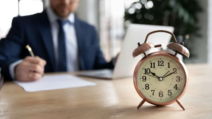 A man in a suit sits at a desk with an alarm clock.