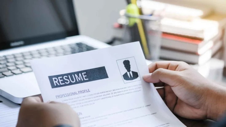 A person holding a resume in front of a laptop.